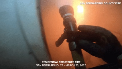 A close-up image taken from a firefighter's helmet camera shows a gloved hand holding a fire hose nozzle inside a smoke-filled structure. The orange glow of active flames can be seen in the background through thick smoke. The environment is intense and low-visibility, highlighting the danger of the scene. The image includes the text: “RESIDENTIAL STRUCTURE FIRE – SAN BERNARDINO, CA – MARCH 23, 2025” and “SAN BERNARDINO COUNTY FIRE” in the upper right corner.